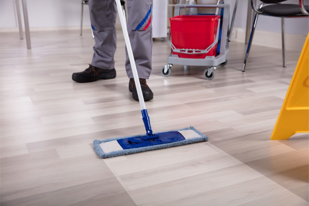 A mop runs over a light hardwood floor, revealing a the clean, light color of the floor under all the dirt and grime.