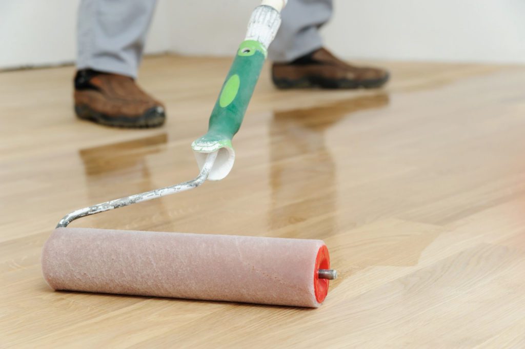 A person applies a hardwood flooring finish with a roller. The refinished areas are shiny and bright.