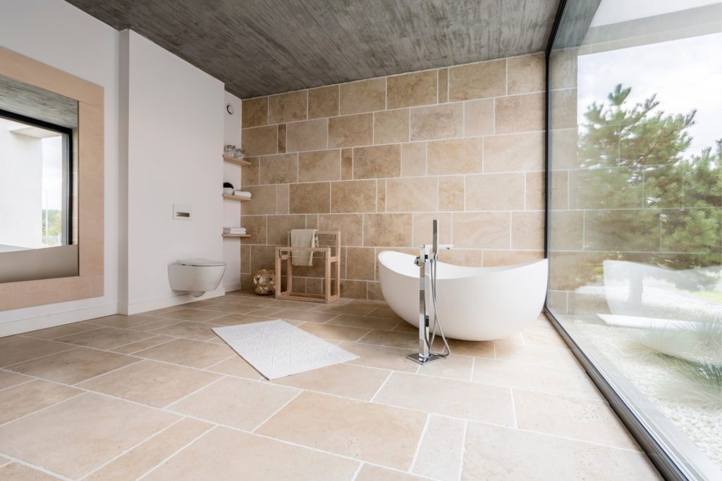 A bright bathroom with travertine bathroom tile on the floor and one accent wall. There is a freestanding tub with modern fixtures and a large window on one wall overlooking an area with trees.