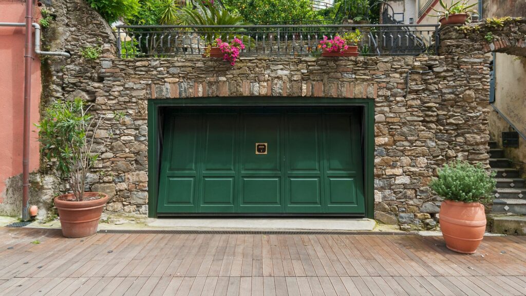 A garage with a green door surrounded by flowers that is going to undergo a garage conversion project 